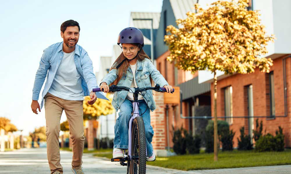 On a nice street, a dad is teaching his daughter to ride a bike